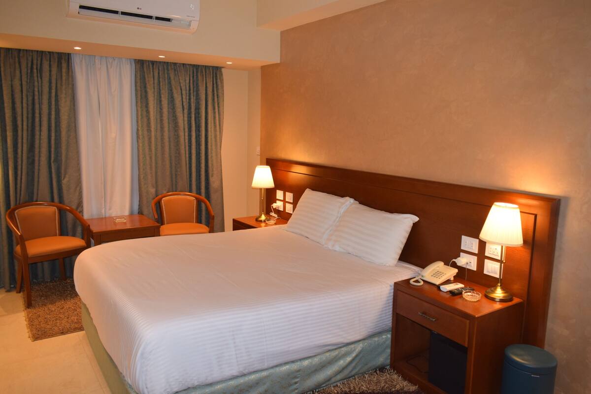 A hotel room with a large bed, two bedside tables with lamps, a desk with a chair, and a window with curtains.