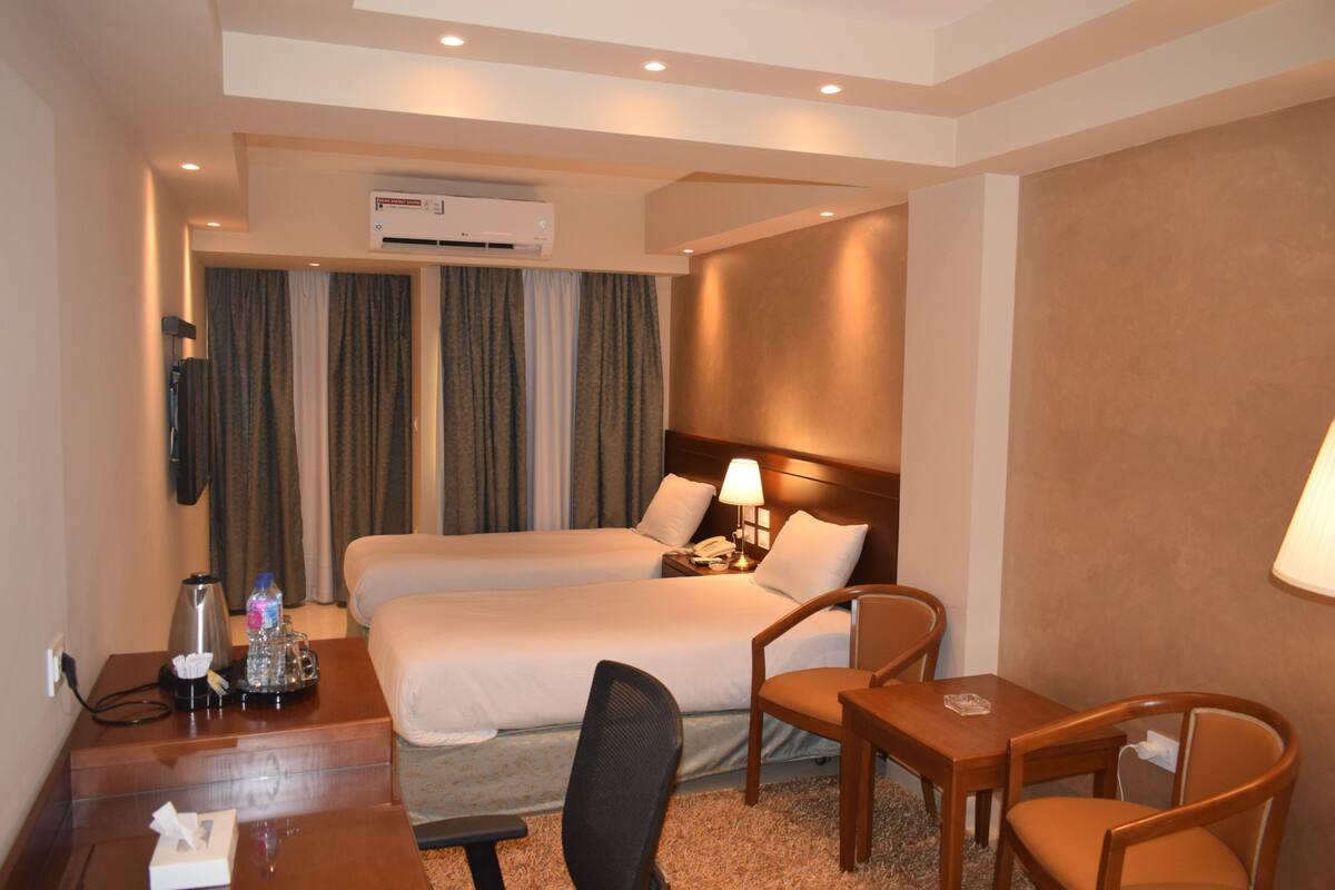 A hotel room with two beds, a desk with a lamp, a chair, a small table, and an air conditioning unit.