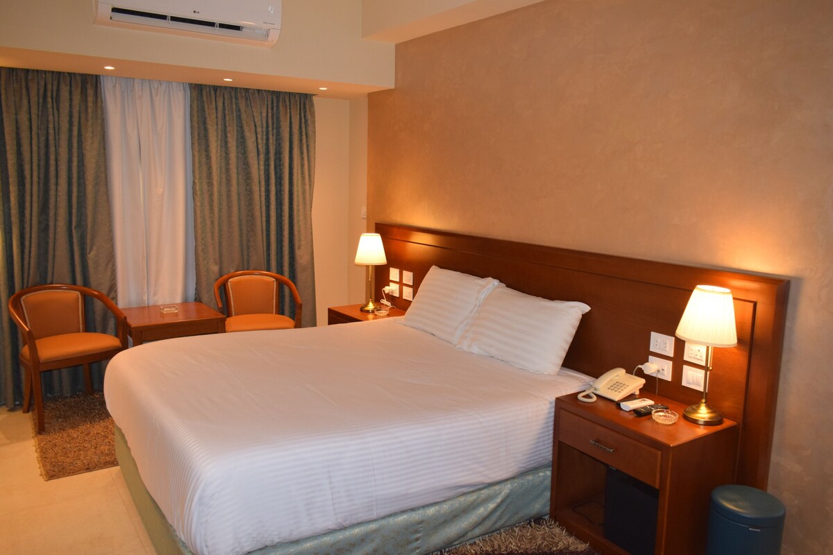 A hotel room with a large bed, two bedside tables with lamps, a desk with a chair, and a window with curtains.