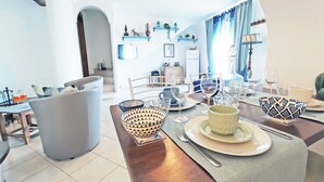 A dining area with a wooden table set for two, a grey armchair, and a sofa in the background. - Estate4home - GOAT PATH (Positano)