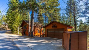 A two-story cabin with a garage, surrounded by tall pine trees. - Exquisite Vacation Home with Hot Tub, EV Charger (Arnold)