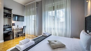 A hotel room with a bed, a desk with a chair, a TV mounted on the wall, and a window with sheer curtains.