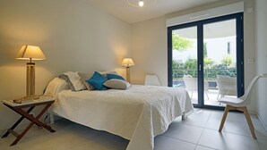 Ein Schlafzimmer mit einem Bett, zwei Nachttischlampen, einem kleinen Tisch, einem Sessel und einer Glastür, die zu einem Außenbereich führt.
