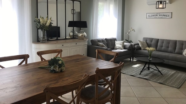A modern living room with a grey sofa, a wooden dining table with chairs, a TV on a cabinet, and a sign reading "Fresh Flower Market".