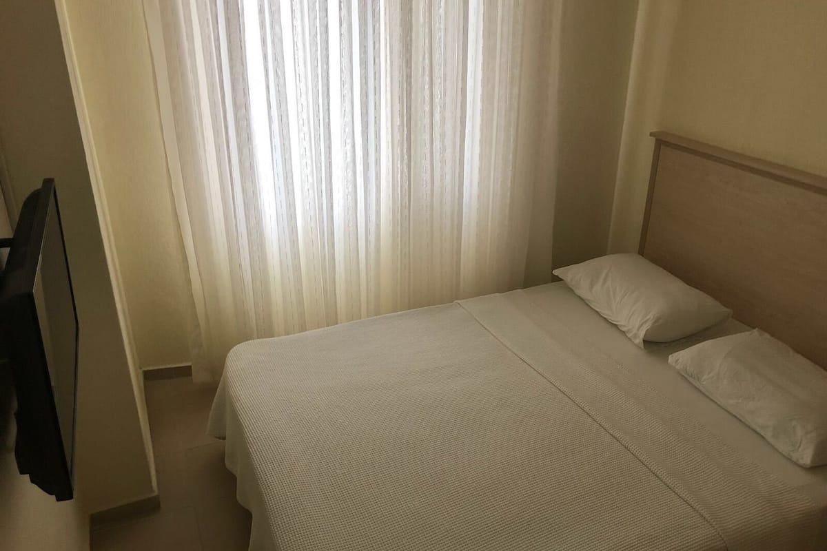 A neatly made bed with white linens in a room with a plain white wall and a window with sheer curtains.
