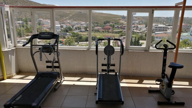 A balcony with exercise equipment including a treadmill, elliptical, and stationary bike, overlooking a residential area with houses and hills. - Elmas Motel (Marmara)