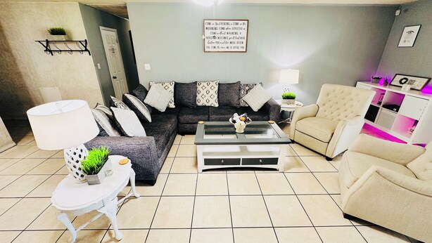 Una sala de estar con un sofá gris, un sillón blanco, una mesa de centro de vidrio y una lámpara. Se observa una pared con una cita enmarcada y una planta pequeña en un estante.
