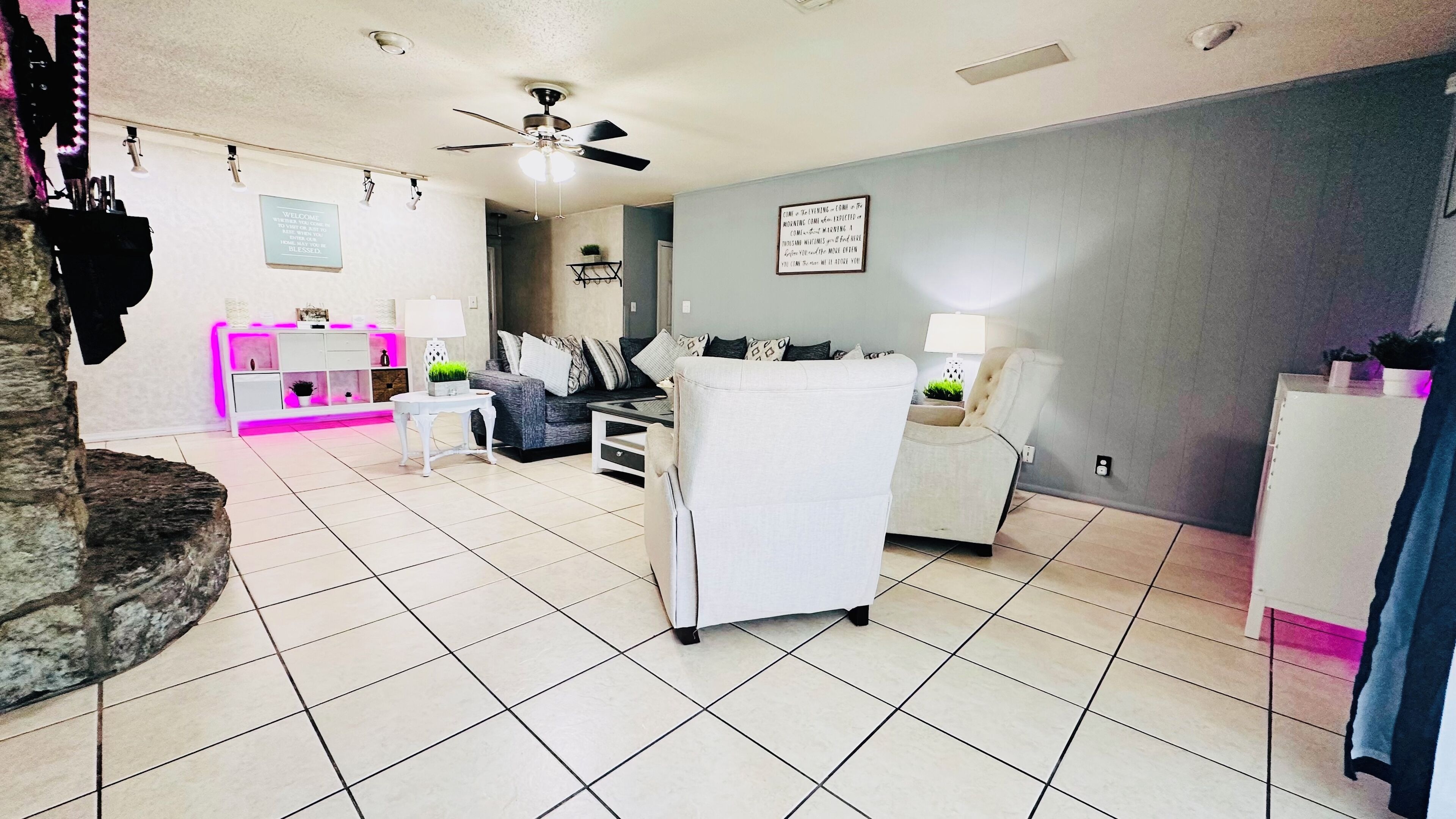 A spacious living room with a grey sofa, a white armchair, and a coffee table. There is a small white side table with a lamp. The room has a tiled floor and a ceiling fan.
