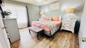 Un dormitorio con cama, mesitas de noche, un banco y una ventana con cortinas.