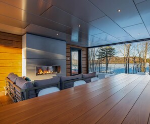 A modern interior with a large wooden table, a fireplace, and a view of a lake and trees.