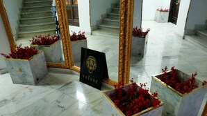 Un vestíbulo con suelo de mármol, una escalera, espejos grandes y arreglos florales rojos en macetas cuadradas.