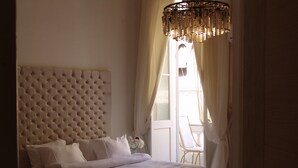 Un dormitorio con cabecera capitonada, cama con ropa de cama blanca, mesita de noche con un florero y una silla junto a la ventana.