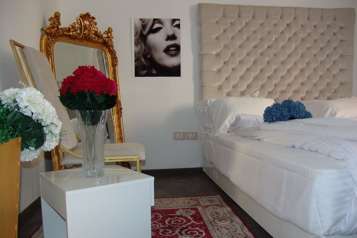 A bedroom with a tufted headboard, a white bed, a red and white patterned rug, a gold-framed mirror, and a vase with flowers.
