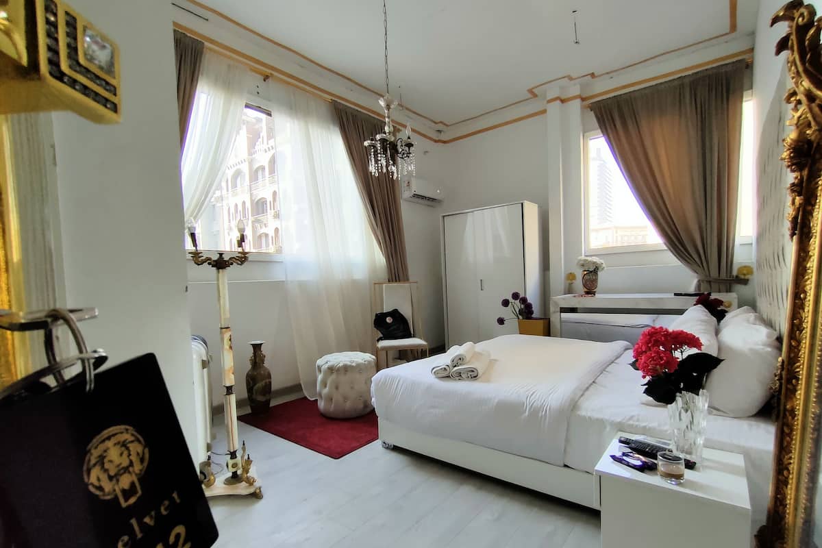 A hotel room with a large bed, a wardrobe, a desk, and a mirror. There is a window with curtains, a chandelier, and a vase with flowers on the desk.