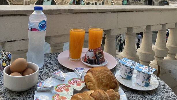 Un desayuno con huevos, jugo, yogur y croissants en un balcón con vistas a edificios históricos.