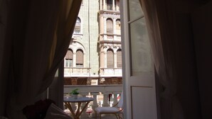 Un balcón con una mesa y sillas, una planta en maceta y vistas a un edificio histórico con ventanas arqueadas y detalles ornamentales.