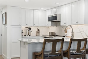 A modern kitchen with white cabinetry, a central island with bar stools, and a stainless steel range hood. - W/D in Unit, Ocean Views Galore, Spectacular Sunrise & Sunset from property (Saint Augustine Beach Florida)