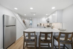 A modern kitchen with white cabinets, a stainless steel refrigerator, a large island with a bowl, and wooden chairs. - W/D in Unit, Ocean Views Galore, Spectacular Sunrise & Sunset from property (Saint Augustine Beach Florida)