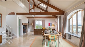 A spacious dining area with a high ceiling and exposed wooden beams, a fireplace, and large windows offering a view of the outdoors. - Oldbury Barn - sleeps 11 guests  in 4 bedrooms (Cheltenham)