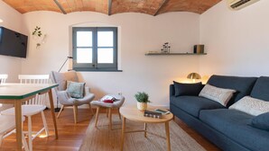 A cozy living room with a wooden table, two chairs, a sofa, and a small coffee table. There is a window with blue shutters, a TV mounted on the wall, and a shelf with books. - Bravissimo Raïms Penthouse, with 2 bedrooms (Girona)