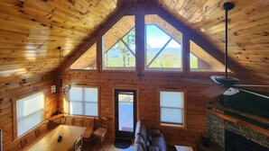 Eine Holzhütte mit geneigtem Dach, einem Essbereich mit Tisch und Stühlen, einem Kamin und einem großen Fenster mit Blick ins Freie.