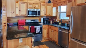 Eine Holzhüttenküche mit einem Edelstahl-Kühlschrank, einer Mikrowelle und einem Ofen. Es gibt Holzschränke, einen Holzbarhocker und ein Fenster mit Blick auf Bäume.