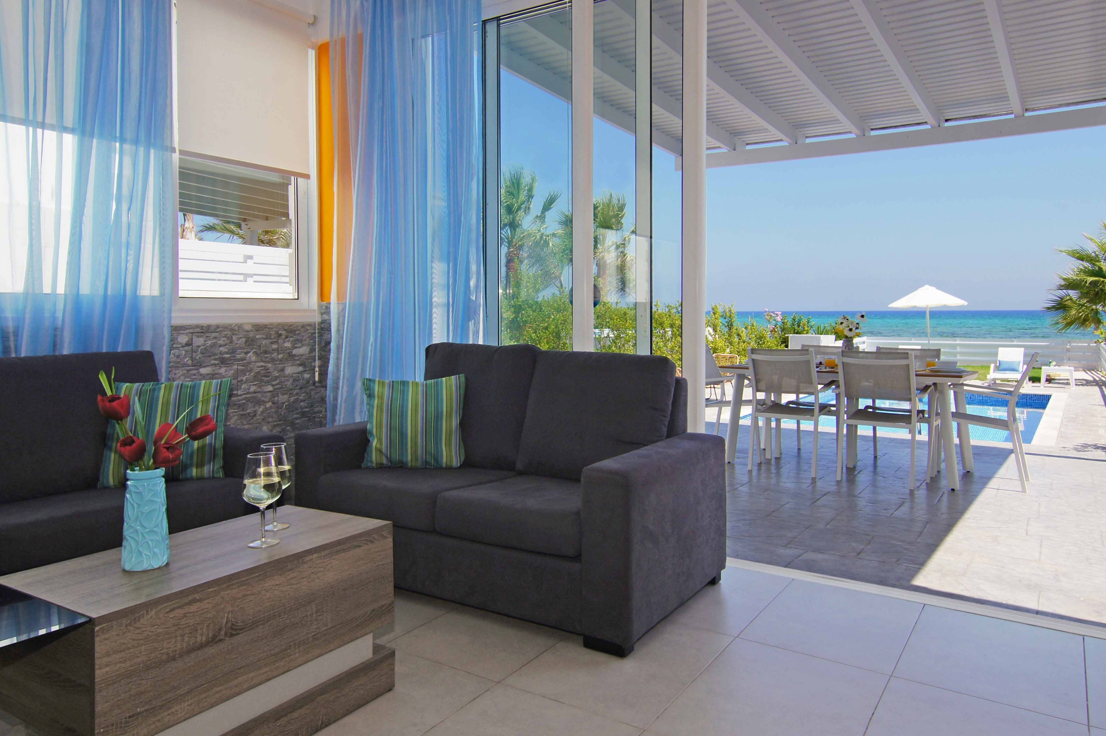 A modern living room with a sofa, coffee table, and a vase of red tulips. There is a large window with blue curtains, and a view of the ocean and a pool area.