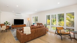 A modern living room with a brown sofa, a TV, and a large window with blinds. - Mirigaan - Pet Friendly - 8 Min Walk to Beach (Culburra Beach)