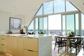 A modern kitchen with a large island, a dining area with a round table, and a panoramic view through large windows. - The Carlton Apartments (Christchurch)
