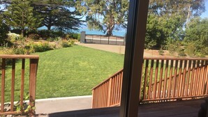 A view from a window overlooking a well-maintained lawn, a wooden deck, and a fenced area with trees and a body of water in the background. - Self contained unit. By the beach and golf course  only 2.2 kms to town centre (Motueka)
