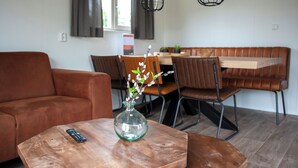 Un salon avec un canapé en cuir marron, une table basse en bois ornée d’un vase et d’une télécommande, et un coin repas comprenant un banc et des chaises en bois.