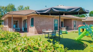 Un edificio de ladrillo con un porche cubierto, mesas al aire libre y una zona de juegos para niños.