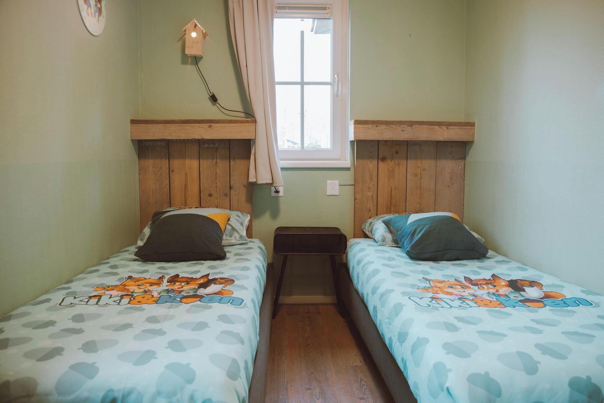 A twin room with two beds, each with a cartoon-themed bedspread, a small wooden nightstand, a wall-mounted lamp, and a window with curtains.