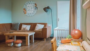 Una sala de estar acogedora con un sofá marrón, una mesa de centro de madera y una pequeña mesa de madera. Hay una decoración en la pared con un zorro y una cita motivacional. Se ve una cuna al fondo.