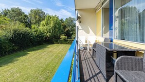 A balcony with wicker furniture, a glass table, and a view of a well-maintained lawn and trees.