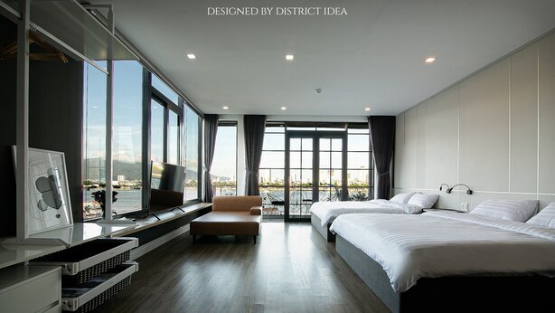 A modern hotel room with a large bed, a sofa, a desk, and a view of the city. - 96 Riverfront Apartment (Đà Nẵng)