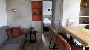 Une chambre moderne avec un canapé gris, une petite table ronde, une table à manger en bois et un lit à l’arrière-plan.