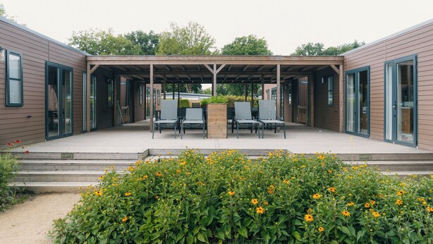 Un espace extérieur aménagé, abrité et doté de tables et de chaises, entouré d’un jardin où poussent des fleurs jaunes.