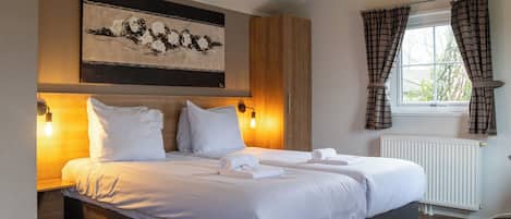 A double bed with white linens, a wooden headboard, a wall-mounted artwork, and a window with plaid curtains.