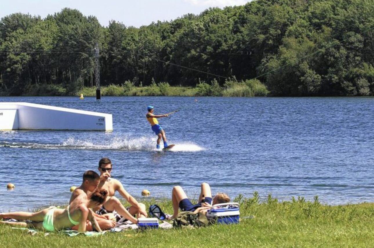 Une scène au bord du lac, avec des personnes pratiquant le wakeboard et d’autres se relaxant sur la rive.