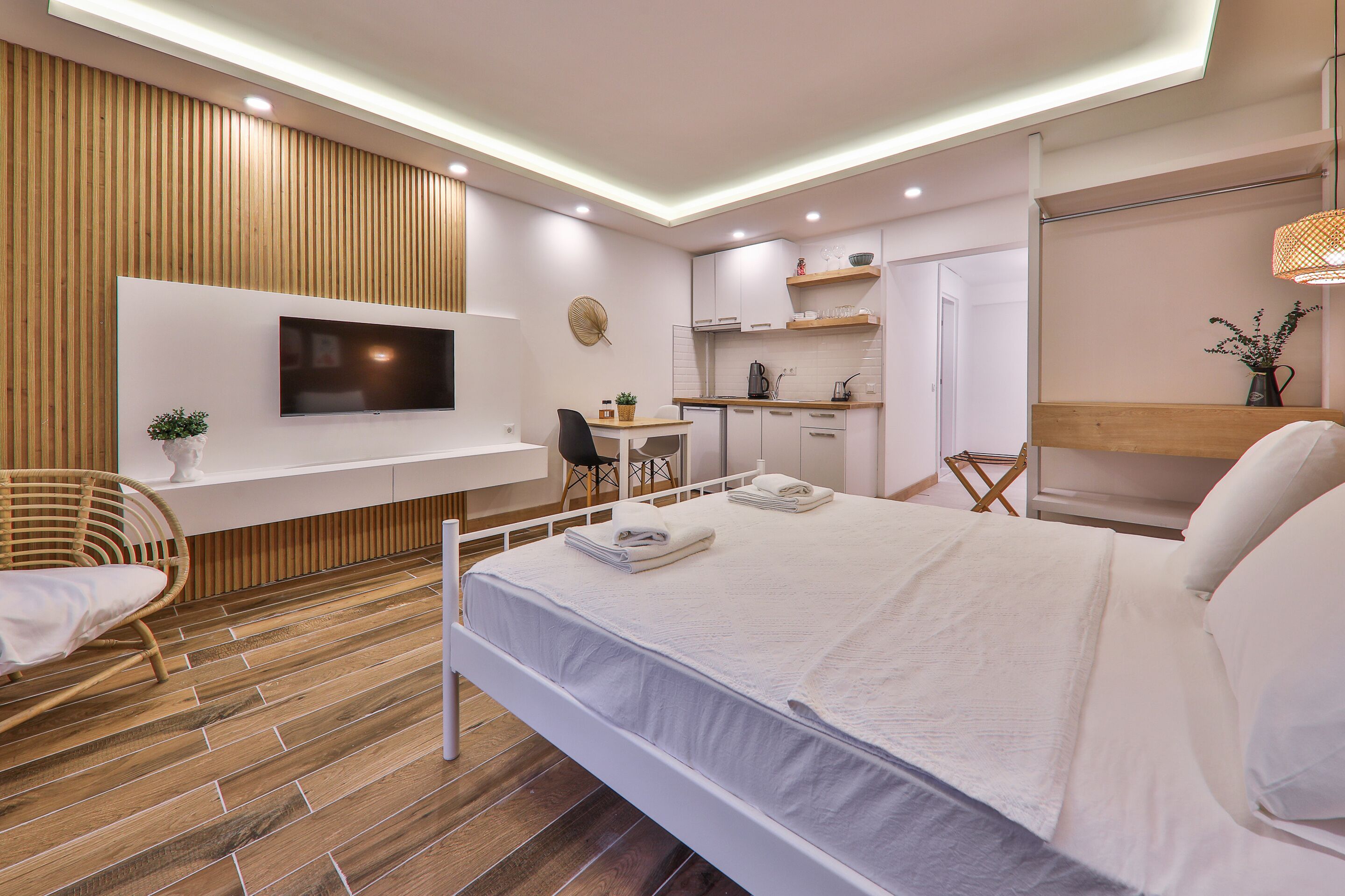 A modern hotel room with a bed, a TV mounted on the wall, a small kitchenette, and a wooden floor.
