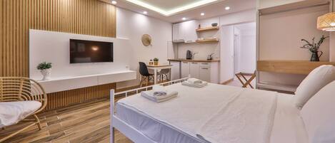A modern hotel room with a bed, a TV mounted on the wall, a small kitchenette, and a wooden floor.