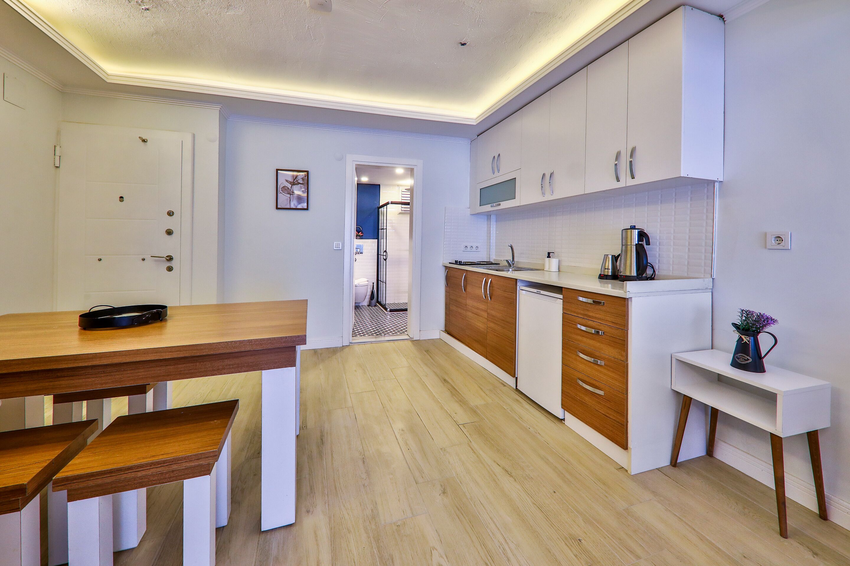 A modern kitchen with white cabinets, wooden countertops, and a built-in oven. There is a dining table with benches and a small white side table with a vase.