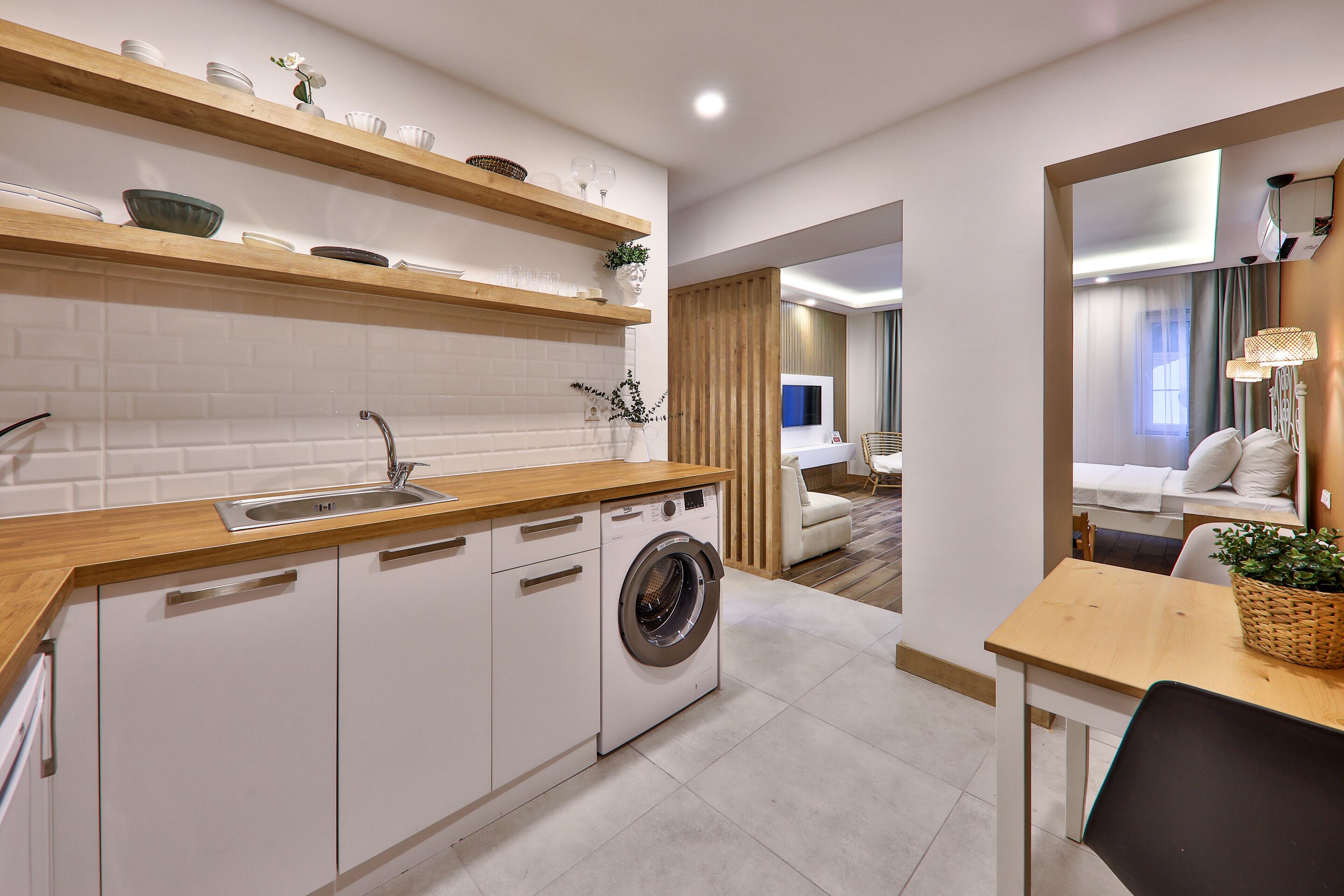 A compact kitchen with a washing machine, wooden shelves, and a dining area with a table and chairs.