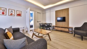 A modern living room with a sofa, coffee table, and TV. There is a wooden wall panel and a dining table with chairs.