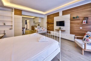 Una habitación de hotel moderna con una cama grande, un televisor montado en la pared y un pequeño rincón de descanso con un banco y un cojín colorido.