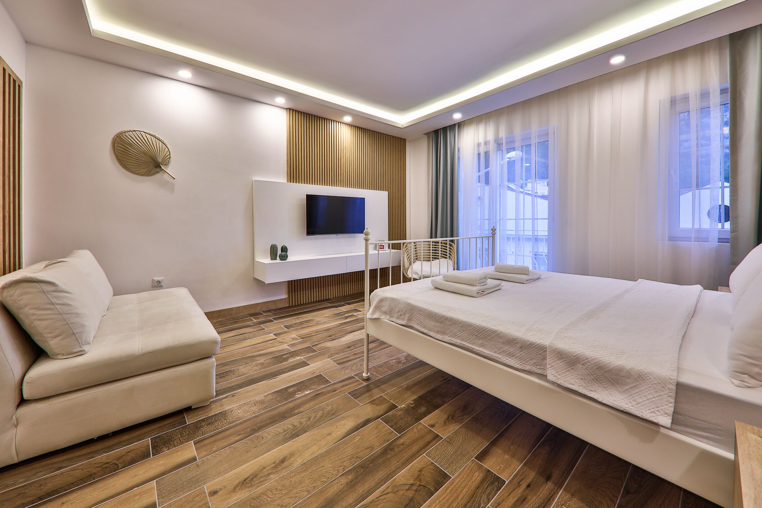 A modern hotel room with a wooden floor, a sofa, a bed with white bedding, a TV mounted on the wall, and a window with curtains.