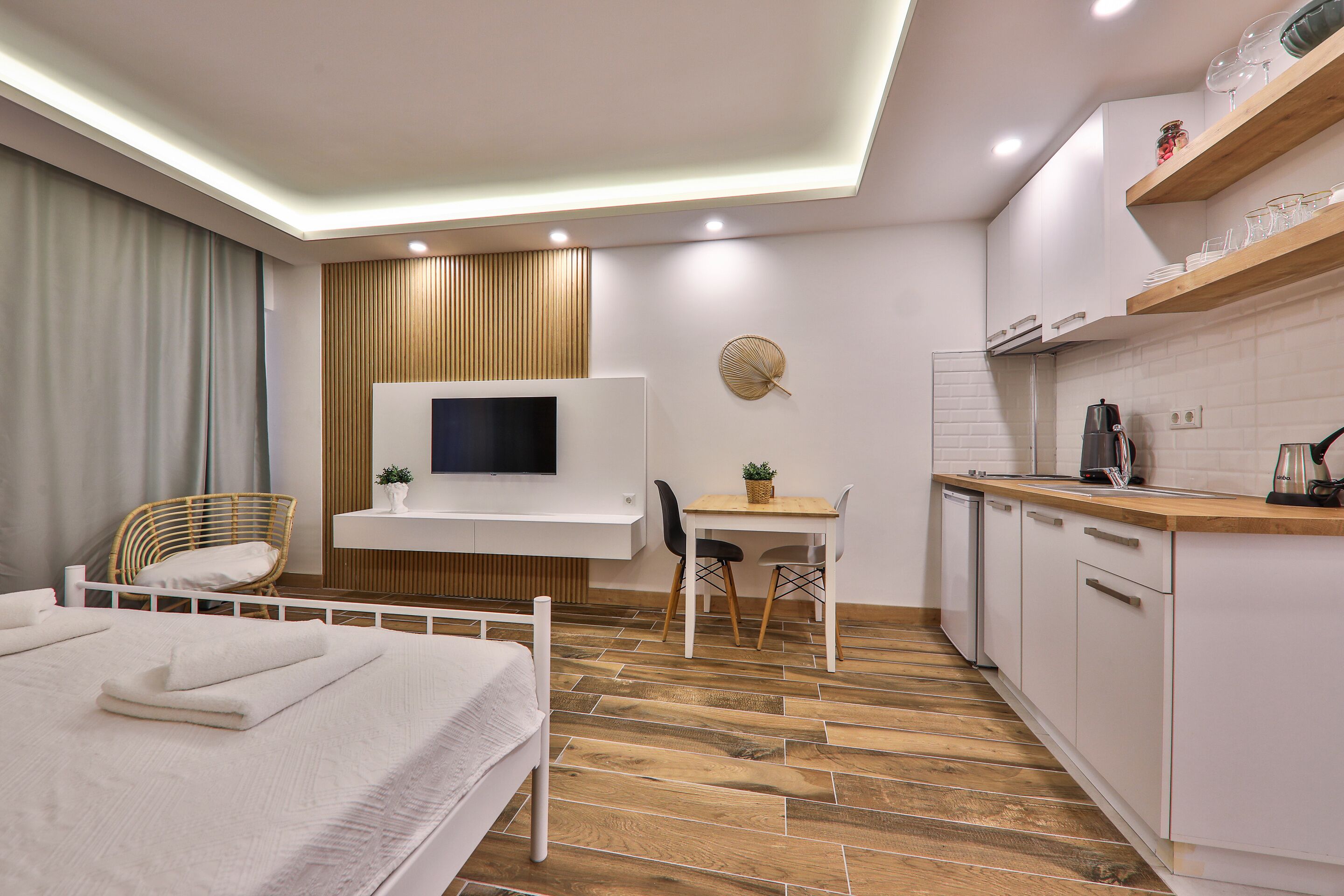 A modern hotel room with a bed, a desk, a TV, and a kitchenette.