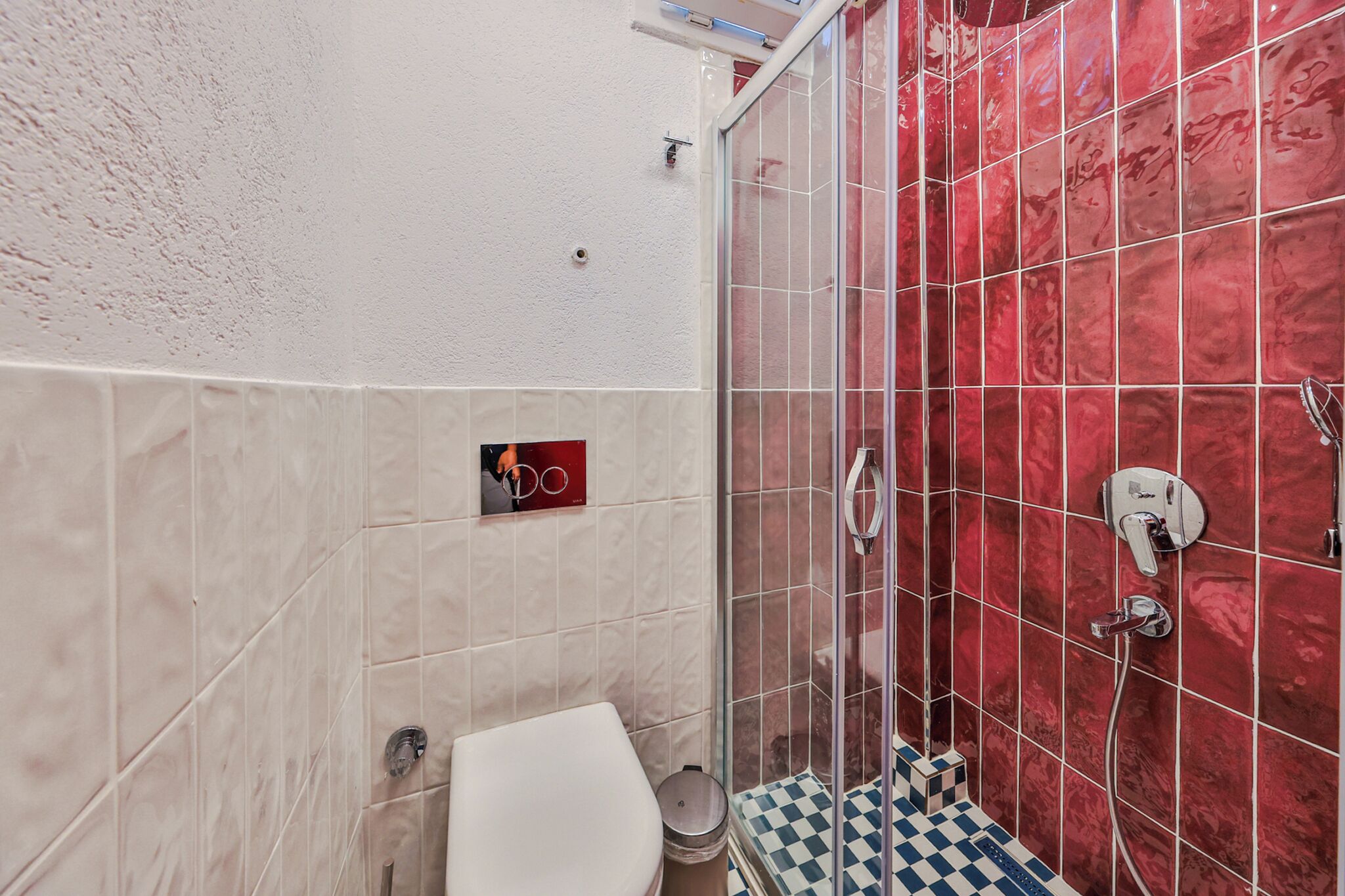 A bathroom with a white toilet, a small trash bin, a shower with red tiles, and a checkered floor.
