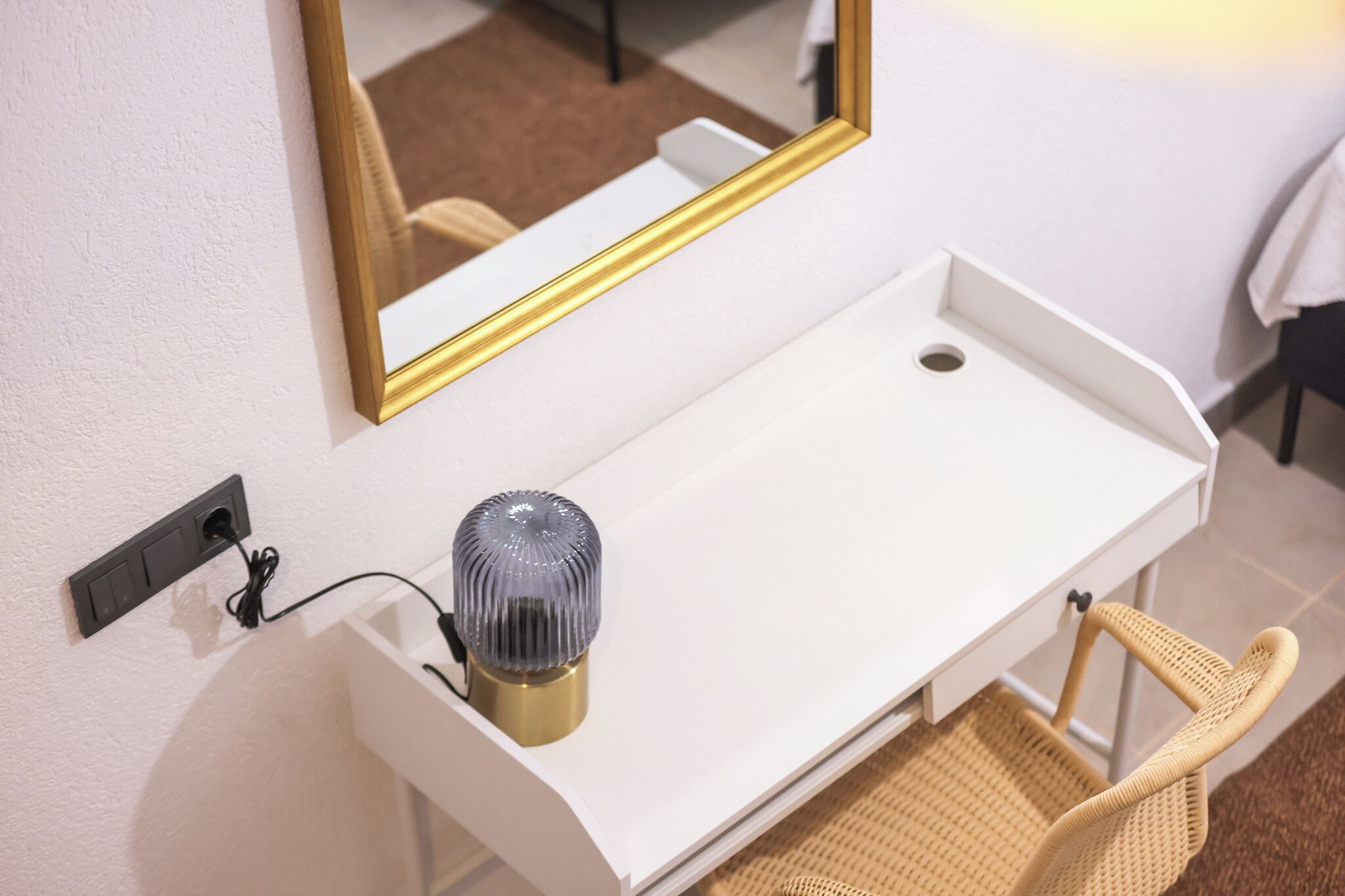 Een witte kaptafel met spiegel, een lamp en een rieten stoel.